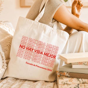 Peut inclure: Sac fourre-tout crème avec le texte rouge "NO HAY VIDA MEJOR" répété. Le texte "Escuela de Precursores 2026" est imprimé en dessous. Le sac, de conception simple, est en tissu résistant, idéal pour les courses ou les livres.