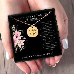 Puede incluir: Un collar con colgante circular dorado grabado con el texto 'no hay vida mejor', presentado en una caja de regalo negra. La caja incluye una ilustraci&oacute;n floral y un texto en espa&ntilde;ol que celebra un bautismo, con la frase '&iexcl;NO HAY VIDA MEJOR!' impresa en el borde inferior del inserto de la caja.