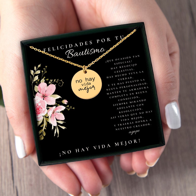Puede incluir: Un collar con colgante circular dorado grabado con el texto 'no hay vida mejor', presentado en una caja de regalo negra. La caja incluye una ilustraci&oacute;n floral y un texto en espa&ntilde;ol que celebra un bautismo, con la frase '&iexcl;NO HAY VIDA MEJOR!' impresa en el borde inferior del inserto de la caja.