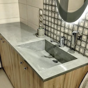 Peut inclure: Un lavabo de salle de bain en béton gris avec un robinet unique, monté sur un meuble-lavabo en bois avec des tiroirs. Le meuble-lavabo est dans une salle de bain avec un mur carrelé et un miroir.