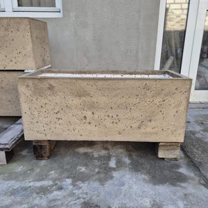 Könnte beinhalten: Ein rechteckiger Betonpflanzkübel mit rauer, strukturierter Oberfläche. Der Pflanzkübel ist hellbraun und steht auf kleinen Holzblöcken. Ein weiterer ähnlicher Pflanzkübel ist im Hintergrund teilweise sichtbar. Der Pflanzkübel ist für den Außenbereich konzipiert.