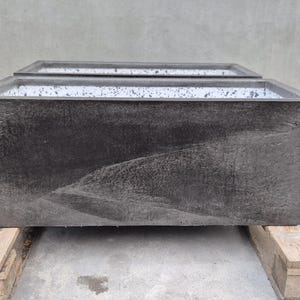 Op de afbeelding: Een rechthoekige plantenbak met een donkergrijze, getextureerde buitenkant en een witte, gespikkelde binnenkant. De plantenbak heeft een brede, open bovenkant en rechte zijkanten, wat zorgt voor een solide en robuuste constructie.