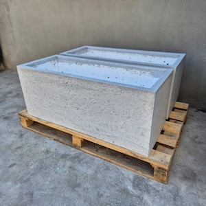 Könnte beinhalten: Zwei rechteckige, hellgraue Betonpflanzgefäße mit strukturierter Oberfläche. Die Pflanzgefäße stehen nebeneinander auf einer Holzpalette. Sie sind für den Außenbereich geeignet.