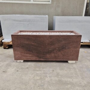 Puede incluir: Una jardinera rectangular de hormigón marrón rojizo con una superficie texturizada. La jardinera está elevada sobre pequeños soportes blancos. En el fondo se ven dos jardineras de hormigón gris adicionales, lo que sugiere un tema de jardín o decoración exterior.
