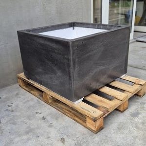 Könnte beinhalten: Ein großer, dunkelgrauer quadratischer Pflanzkübel mit strukturierter Oberfläche. Der Pflanzkübel steht auf einer Holzpalette. Das Innere des Pflanzkübels ist weiß. Der Pflanzkübel besteht aus einem festen Material.
