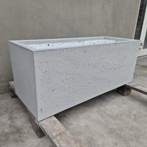 Könnte beinhalten: Ein rechteckiger Pflanzkübel aus Beton mit einer hellgrauen, porösen und strukturierten Oberfläche. Der Pflanzkübel verfügt über einen hohlen Innenraum mit einer glatten, hellgrauen Auskleidung und scharfen, klaren Kanten.