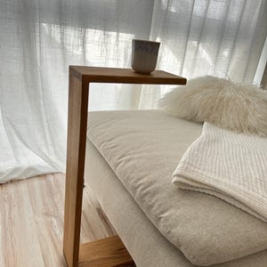 Puede incluir: Una mesa auxiliar de madera con un diseño en forma de C. La mesa tiene una parte superior rectangular y una sola pata que se curva para formar una base. Una pequeña taza de cerámica está sobre la mesa. La mesa está junto a un sofá beige con una manta y una almohada blancas.