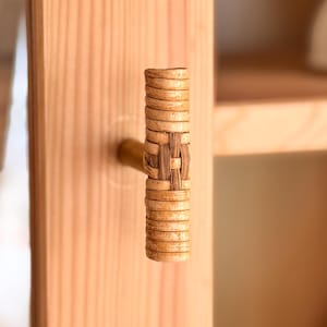 May include: Close-up of a wooden cabinet door with a woven rattan handle. The handle is cylindrical with a woven center section and is attached to a light-colored wooden door. The cabinet is part of a larger wooden storage unit.