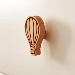 May include: A light brown wooden drawer knob shaped like a hot air balloon. The front surface features engraved vertical lines and a basket detail, creating a textured silhouette. The knob is mounted onto a smooth, off-white flat surface.