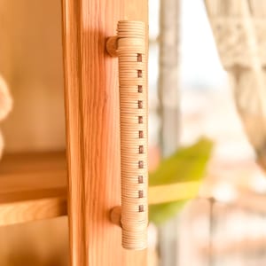 Può includere: Primo piano di un'anta di un armadio in legno con una maniglia cilindrica. La maniglia è realizzata in legno chiaro con un design strutturato e scanalato e piccoli accenti rettangolari. L'anta dell'armadio è anch'essa in legno chiaro.