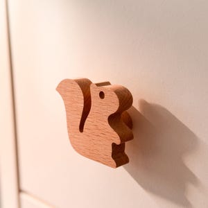 Op de afbeelding: Een lichtbruine houten meubelknop in de vorm van een eekhoorn, met een gladde afwerking en een gedetailleerd silhouet. De knop is bevestigd aan een platte, crèmekleurige ladefront en werpt een duidelijke schaduw naar de rechterkant.