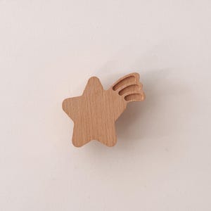 May include: A light brown wooden drawer knob shaped like a shooting star with a smooth finish. The star features a central body with three trailing lines extending from the upper right side, attached to a flat, solid white surface panel.