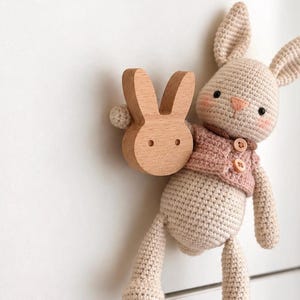May include: A beige crocheted rabbit doll wearing a dusty pink vest with two small buttons, holding a smooth, light-colored wooden rabbit-shaped knob. The rabbit has embroidered eyes, a pink nose, and pink cheeks, attached to a white drawer.