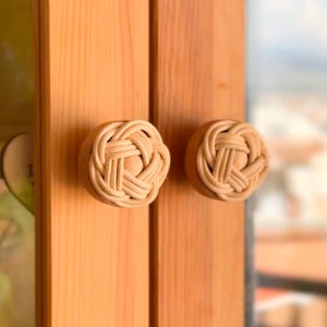 May include: Two wooden door knobs with a woven, basket-like design. The knobs are light beige and are mounted on a light brown wooden door. The door has a natural wood grain and is part of a set of double doors.