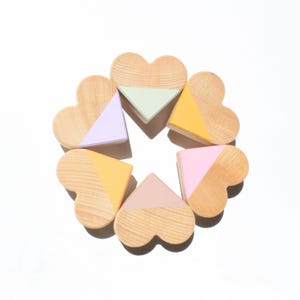 May include: Six heart-shaped wooden pieces arranged in a circular pattern. Each piece features a natural wood grain finish on the top half and a solid matte colour on the bottom half, including shades of light purple, mint green, yellow, pink, and peach.