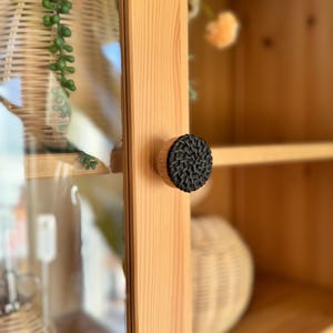 May include: A close-up of a black woven knob on a wooden cabinet door. The knob is round and has a slightly raised design. The cabinet door is made of light wood and has a glass panel.