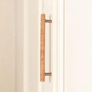 May include: A vertical door pull with a woven rattan center and bronze-colored metal mounting hardware. The door pull is a light tan color and is mounted on a white door. The hardware is a simple, modern design.