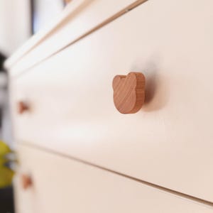 Può includere: Una cassettiera color beige chiaro con diversi cassetti, ognuno dotato di un pomello in legno liscio color marrone chiaro a forma di testa di orso. I pomelli a forma di orso sono fissati al centro dei frontali dei cassetti come maniglia.