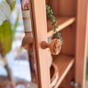 May include: Close-up of a wooden cabinet door with a decorative knob. The knob features a round, woven design in a light brown color, contrasting with the smooth, light-colored wood of the door. The cabinet has shelves and a glass door.