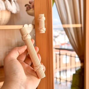 May include: A light-colored wooden cabinet door handle with a woven design. The handle is cylindrical with a series of vertical wooden rods held together by woven knots at each end. A matching handle is attached to the cabinet door.