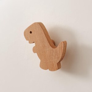 May include: A light brown wooden drawer knob shaped like a dinosaur. The knob features a smooth, natural wood grain finish and a simple silhouette of a standing dinosaur with a small circular eye detail, attached to a circular mounting base.