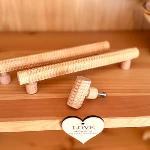 Op de afbeelding: Drie geweven rotan kastknoppen met een houten basis. De knoppen zijn in verschillende maten en worden getoond op een houten oppervlak. Een hartvormig bord met de tekst "LOVE by HANDEYAPIMI" is op de voorgrond.