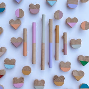 Può includere: Una collezione di pomelli e maniglie in legno per mobili, inclusi tiratori cilindrici lunghi e varie forme come cerchi, cuori e teste d'orso. Ogni pezzo presenta una finitura in legno naturale abbinata a sezioni dipinte in colori pastello.