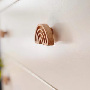 Può includere: Pomello per cassetto a forma di arcobaleno in legno. Il pomello è realizzato in legno chiaro e presenta un design ad arcobaleno intagliato. Il pomello è attaccato a una porta di un armadio bianco. Il design ad arcobaleno aggiunge un tocco stravagante.