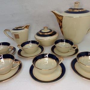 Pode incluir: Um conjunto de chá vintage creme com detalhes em azul marinho e dourado. O conjunto inclui uma chaleira, açucareiro, cremeira e várias chávenas de chá com pires. O conjunto de chá é provavelmente feito de porcelana.