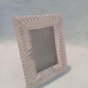 Può includere: Una cornice rettangolare rosa chiaro con un design a corda intrecciata sui bordi. La cornice è realizzata in un materiale traslucido ed è vuota. La cornice è leggermente angolata, pronta per contenere una fotografia.