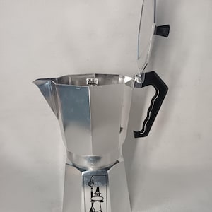 May include: A silver stovetop espresso maker with a black handle and lid. The lid is open, revealing the interior. A small graphic of a man with a hat is on the front. The coffee pot is made of metal.