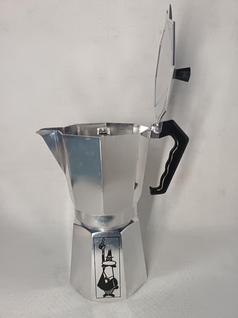 May include: A silver stovetop espresso maker with a black handle and lid. The lid is open, revealing the interior. A small graphic of a man with a hat is on the front. The coffee pot is made of metal.