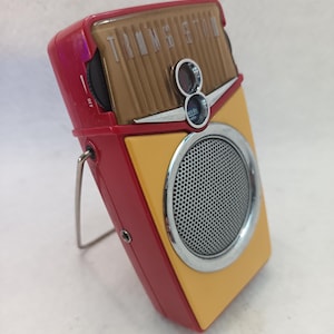 Könnte beinhalten: Ein Retro-Transistorradio in Rot, Gelb und Braun. Das Radio verfügt über einen Lautsprecher mit silberner Umrandung, einen Abstimmknopf und das Wort "TRANSISTOR" auf der Oberseite. Es hat einen Metallständer und einen "OFF"-Schalter.