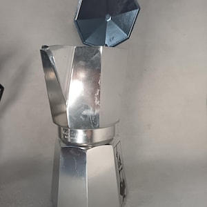 May include: A silver stovetop espresso maker with a detached lid. The coffee maker has a classic octagonal design and is made of shiny metal. The lid is also silver and has a handle. The coffee maker is designed to brew espresso.
