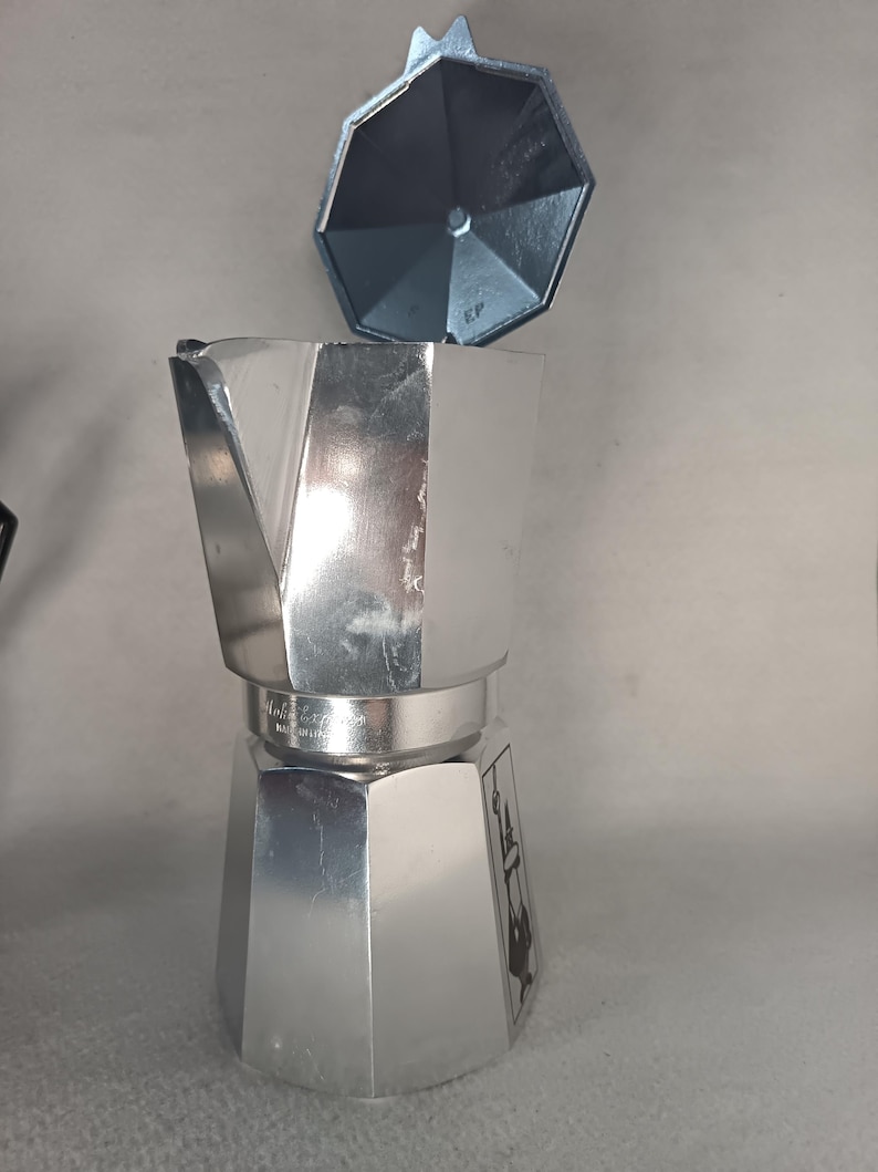 May include: A silver stovetop espresso maker with a detached lid. The coffee maker has a classic octagonal design and is made of shiny metal. The lid is also silver and has a handle. The coffee maker is designed to brew espresso.