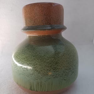 Op de afbeelding: Een keramische vaas met een afgeronde body en een cilindrische bovenkant. De vaas heeft een gespikkelde groene glazuur over een bruine basis, met een bruine bovenkant. Het ontwerp is eenvoudig en rustiek, geschikt voor woondecoratie.