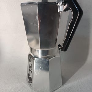 May include: A silver stovetop espresso maker with a black handle and lid knob. The coffee maker has a classic octagonal design and a small graphic on the front. This Italian-style coffee maker is designed for brewing espresso on a stovetop.