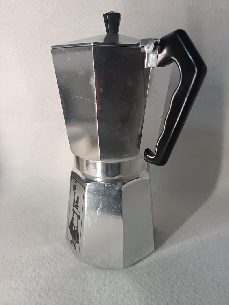 May include: A silver stovetop espresso maker with a black handle and lid knob. The coffee maker has a classic octagonal design and a small graphic on the front. This Italian-style coffee maker is designed for brewing espresso on a stovetop.
