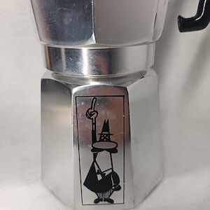May include: A silver stovetop espresso maker with a black handle. The coffee pot features a black and white cartoon figure on the front. The figure is wearing a hat and pointing upwards. The espresso maker is made of metal.