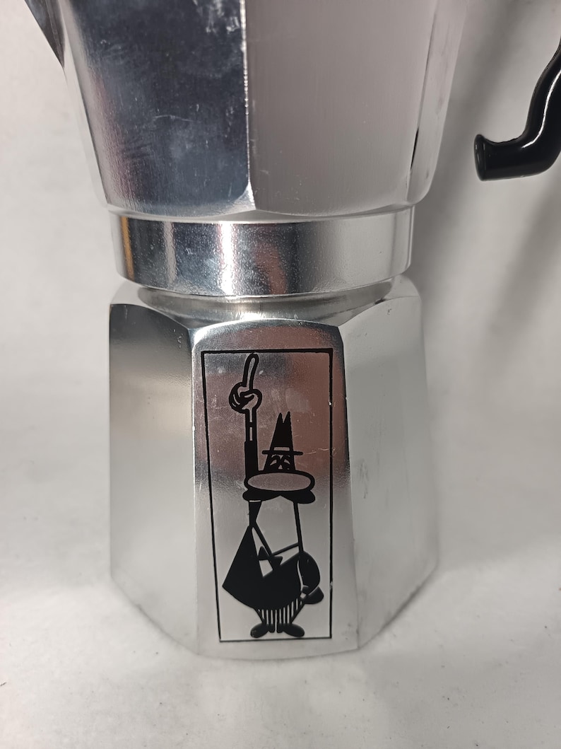 May include: A silver stovetop espresso maker with a black handle. The coffee pot features a black and white cartoon figure on the front. The figure is wearing a hat and pointing upwards. The espresso maker is made of metal.