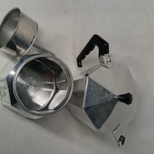 May include: A disassembled silver stovetop espresso maker with a black handle. The coffee maker is made of shiny metal and has a geometric design. The top part is separated from the base, showing the inside.