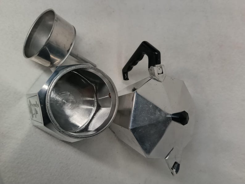 May include: A disassembled silver stovetop espresso maker with a black handle. The coffee maker is made of shiny metal and has a geometric design. The top part is separated from the base, showing the inside.