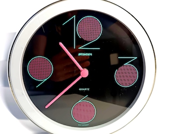 May include: A round wall clock with a white frame and a black face. The dial features large teal numerals 12, 3, 6, and 9, each containing a pink dotted circle. The clock has pink hour and minute hands, with the text Staiger and Quartz printed in teal, and West Germany printed at the bottom.