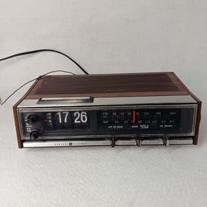 Può includere: Una radiosveglia AM/FM vintage General Electric con involucro in plastica marrone e pannello frontale argentato. Il dispositivo presenta un orologio digitale meccanico a palette che segna le 17:26, un sintonizzatore radio orizzontale e manopole.