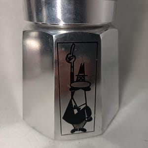 May include: A silver-colored stovetop espresso maker with a black handle. The coffee maker features a black and white cartoon illustration on the front. The design is a classic Italian coffee maker.