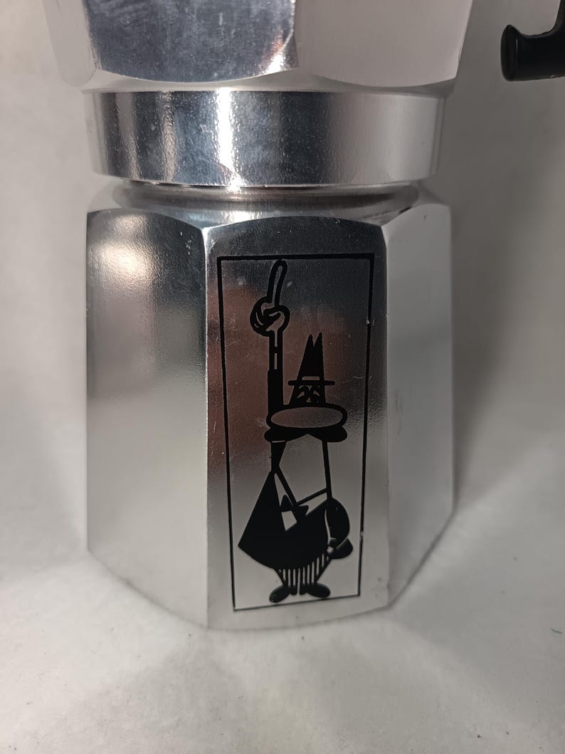 May include: A silver-colored stovetop espresso maker with a black handle. The coffee maker features a black and white cartoon illustration on the front. The design is a classic Italian coffee maker.
