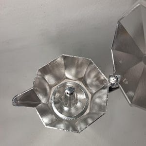 May include: A silver stovetop espresso maker, viewed from above, with the lid open. The pot has a faceted design and a spout. The interior is visible, showing the filter basket and the central column. The lid is hinged and has a circular design.
