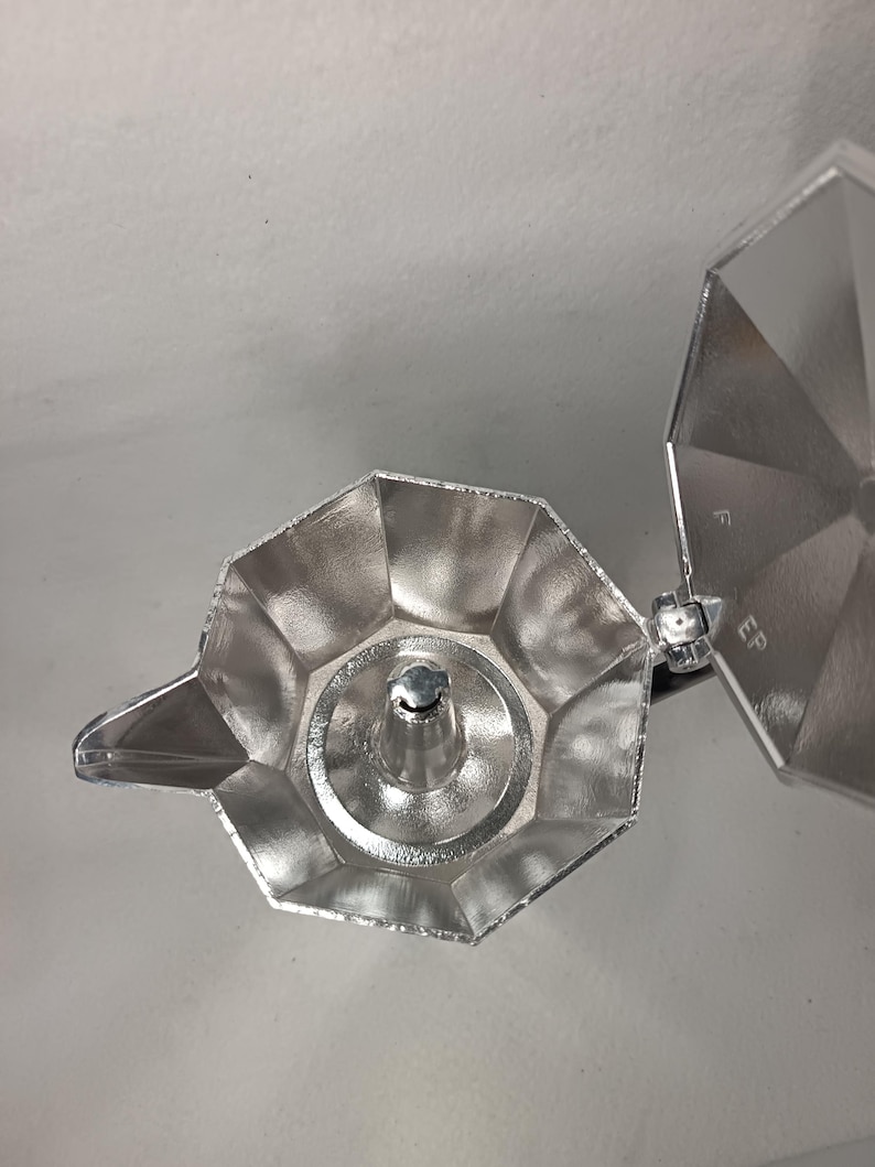 May include: A silver stovetop espresso maker, viewed from above, with the lid open. The pot has a faceted design and a spout. The interior is visible, showing the filter basket and the central column. The lid is hinged and has a circular design.