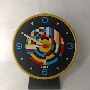 Puede incluir: Un reloj Brio vintage con esfera negra, marco amarillo y diseño geométrico colorido. El reloj tiene manecillas blancas, marcadores de hora triangulares negros y la palabra "BRIO" impresa en la esfera. Está montado sobre una base negra.