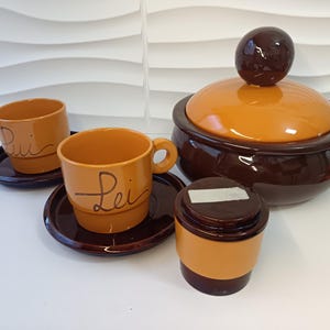 May include: A set of brown and orange ceramic teacups and saucers with the words "Lui" and "Lei" written on them. The set also includes a brown and orange ceramic sugar bowl with a lid.